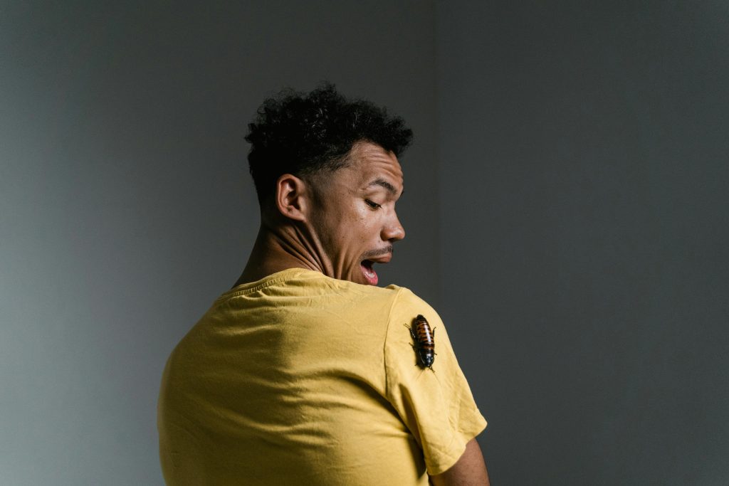 A man in a yellow shirt looks scared as a cockroach sits on his shoulder.